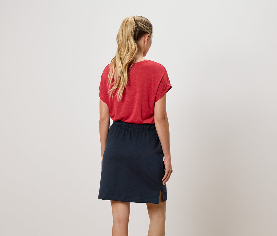 Vue de dos d'une femme avec une queue de cheval blonde portant un t-shirt rouge et une jupe bleu marine.