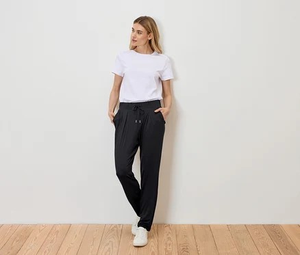 Une femme se tient debout, les mains dans les poches, devant un mur, portant un t-shirt blanc, un pantalon noir à cordon de serrage et des baskets blanches.