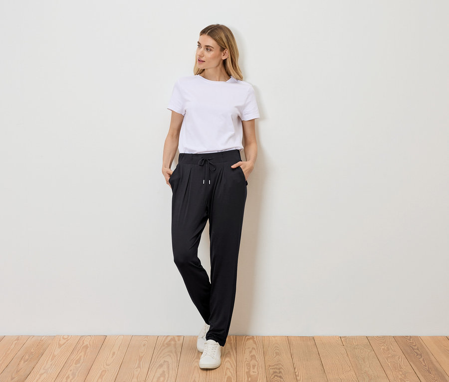 Une femme se tient debout, les mains dans les poches, devant un mur, portant un t-shirt blanc, un pantalon noir à cordon de serrage et des baskets blanches.