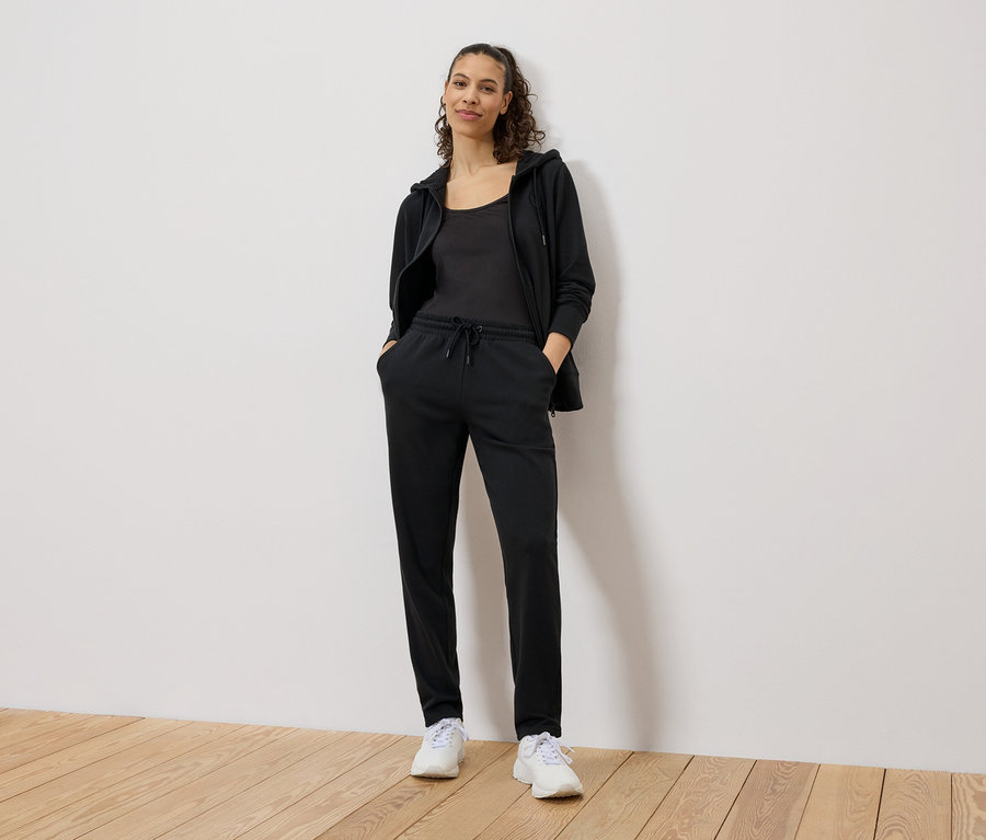 Une femme se tient debout, les mains dans les poches d'un pantalon de jogging noir, portant un débardeur noir, un sweat à capuche noir et des baskets blanches.