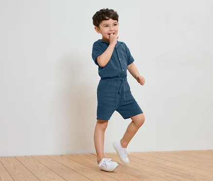 Un petit garçon en combinaison de coton bleue et baskets blanches se tient sur un plancher en bois et suce son doigt.