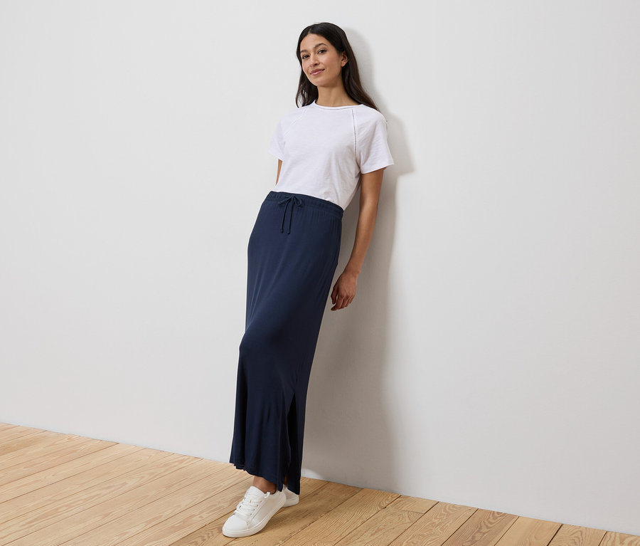 Une femme est appuyée contre un mur, portant un t-shirt blanc, une longue jupe bleu marine et des baskets blanches.