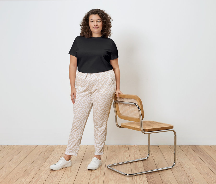 Une femme aux cheveux bouclés se tient à côté d'une chaise, portant un t-shirt noir, un pantalon à motifs et des baskets blanches.