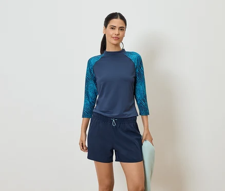 Une femme portant un haut bleu marine à manches trois-quarts et un short bleu tient une serviette bleu clair devant un fond blanc.