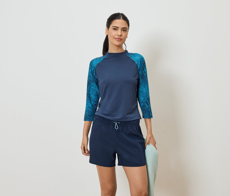 Une femme portant un haut bleu marine à manches trois-quarts et un short bleu tient une serviette bleu clair devant un fond blanc.