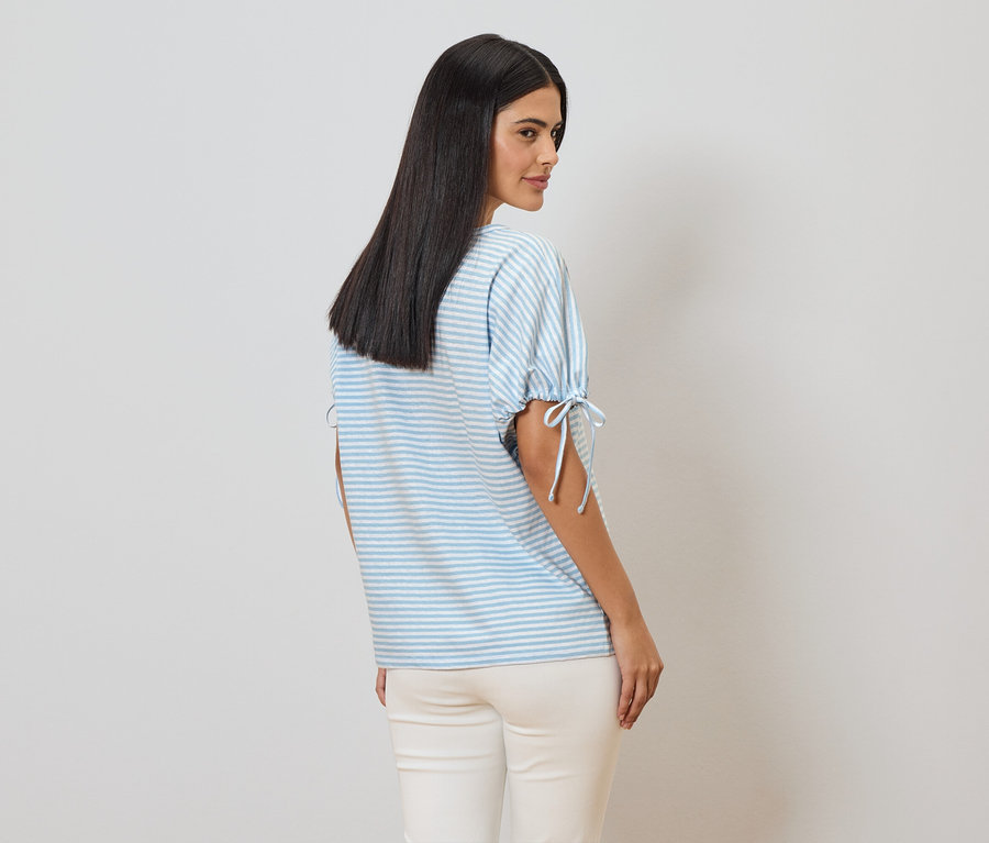 Une femme aux longs cheveux foncés porte un t-shirt rayé bleu clair et blanc avec des liens aux manches et un pantalon clair.