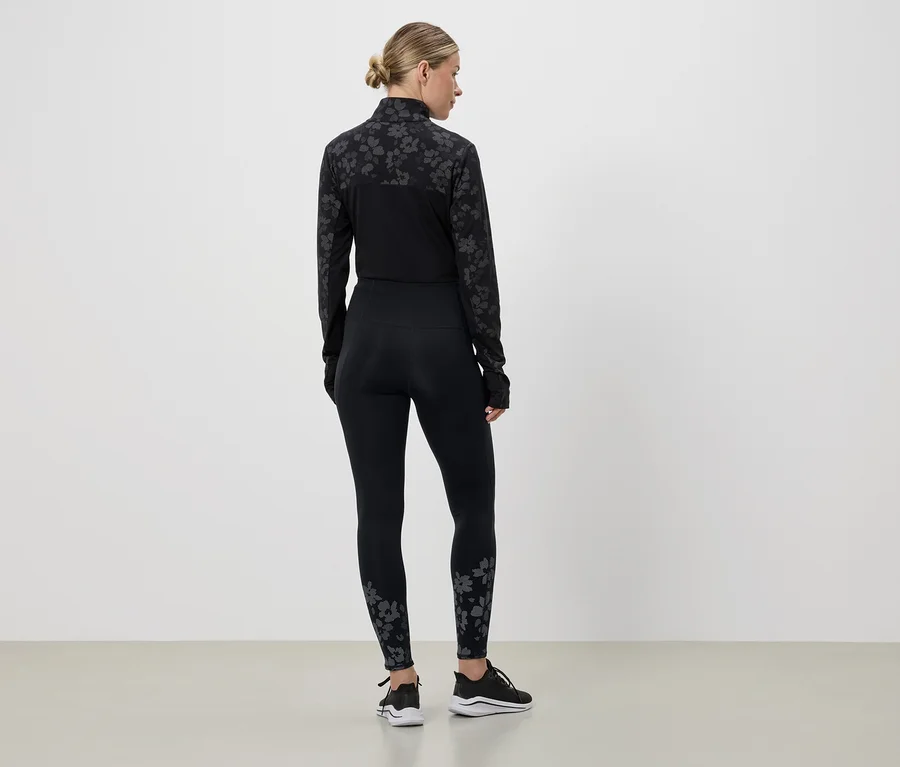 Vue arrière d'une femme portant un survêtement de sport noir à motif floral, des baskets noires et les cheveux attachés en queue de cheval, devant un mur blanc.