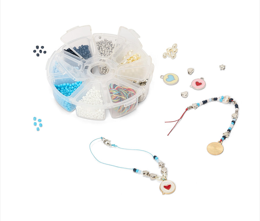 Boîte ronde ouverte contenant diverses perles, élastiques et breloques. Deux bracelets avec des breloques en forme de cœur sont à côté.