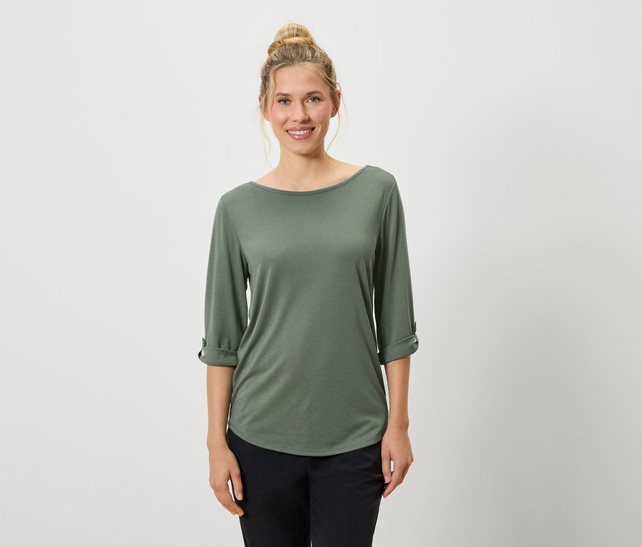 Une femme aux cheveux blonds relevés porte un T-shirt olive à manches longues et un pantalon noir sur un fond blanc.