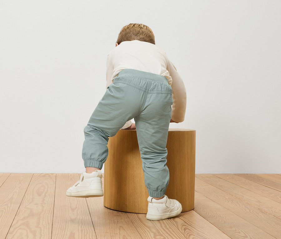 Un enfant grimpe sur un tabouret en bois, portant un haut à manches longues et un pantalon bleu clair.