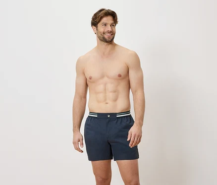 Un homme torse nu porte un caleçon boxer bleu avec une ceinture blanche et verte sur fond blanc.