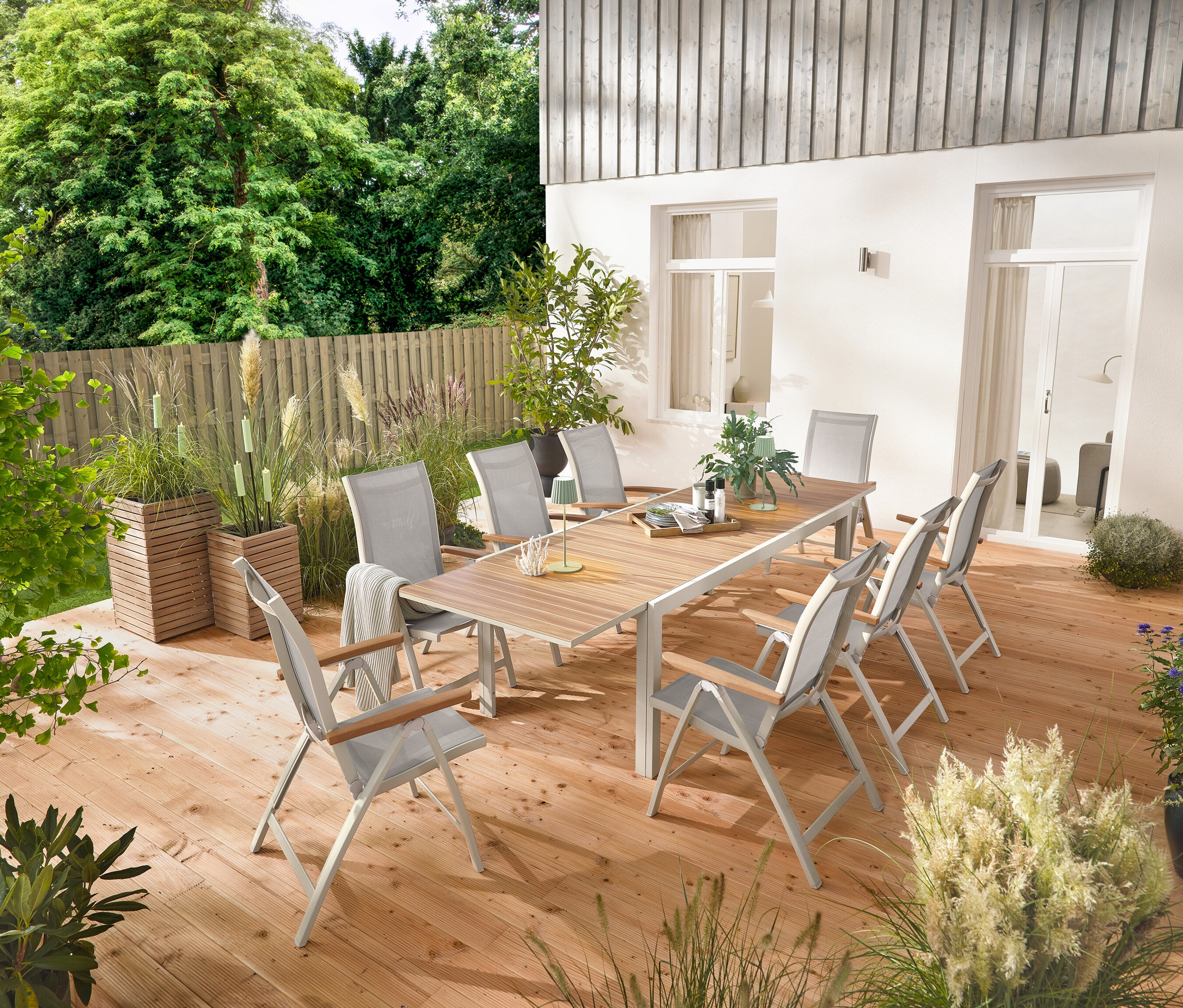 Une table de jardin extensible en aluminium « Liska » d'environ 200 à 300 cm de long est installée sur une terrasse en bois, entourée de six fauteuils à dossier haut « Liska » avec dossier réglable. Deux colonnes à plantes « Tinus » et cinq torches de jardin à LED sont également visibles.