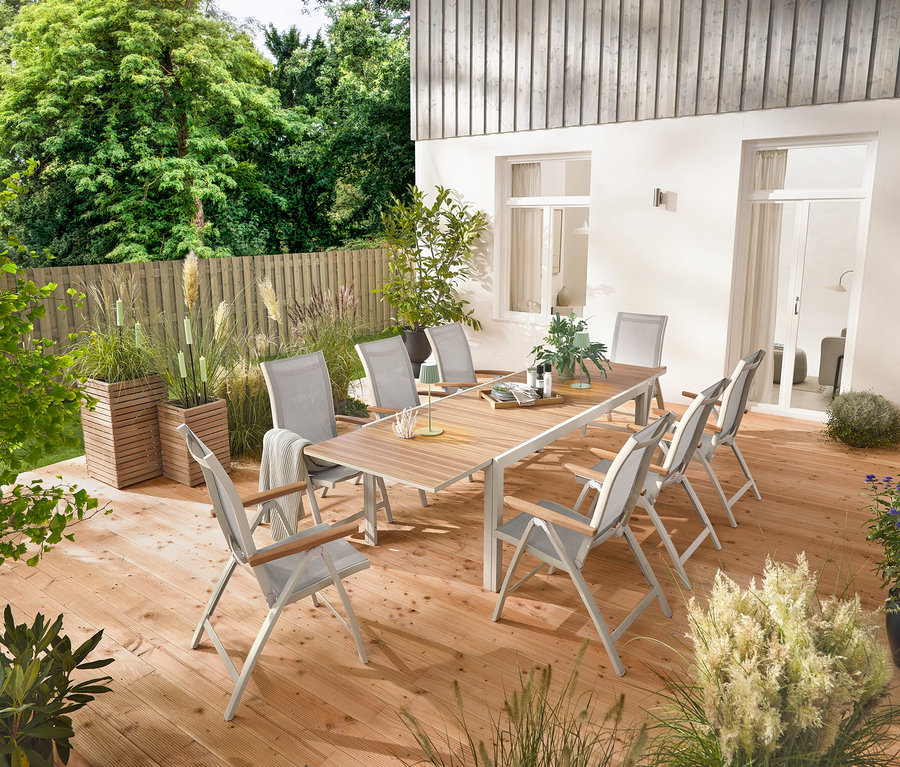 Une table de jardin extensible en aluminium « Liska » d'environ 200 à 300 cm de long est installée sur une terrasse en bois, entourée de six fauteuils à dossier haut « Liska » avec dossier réglable. Deux colonnes à plantes « Tinus » et cinq torches de jardin à LED sont également visibles.