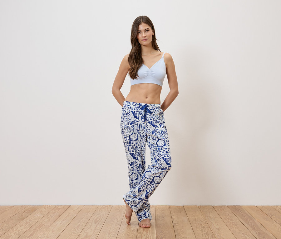 Une femme portant un soutien-gorge bleu clair et un pantalon de pyjama à motifs bleus et blancs se tient devant un mur blanc.