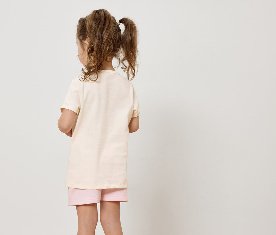 Une fille aux cheveux blonds portant un t-shirt beige et un short rose se tient dos à la caméra sur un fond blanc.