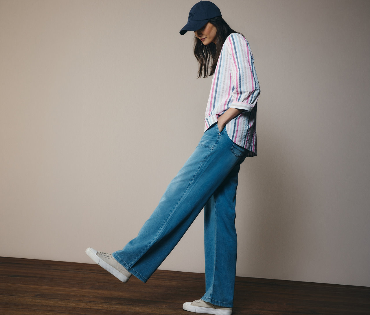 Une femme portant un jean et une casquette de baseball porte un chemisier CECIL rayé. Elle se tient sur un plancher de bois.