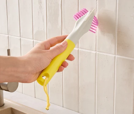 Une main tient une brosse jaune et grise à poils roses et nettoie les joints de carrelage.