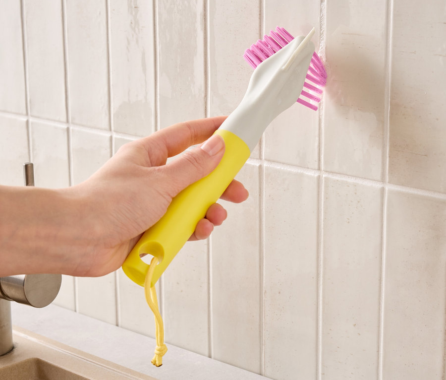 Une main tient une brosse jaune et grise à poils roses et nettoie les joints de carrelage.