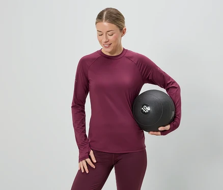 Une femme en tenue de sport bordeaux tient un médecine-ball noir de 10 kg sur un fond gris.