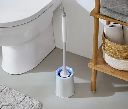 Dans la salle de bain, des toilettes blanches sont à côté d'une brosse WC avec une lumière bleue et d'une étagère en bois avec des serviettes et du papier toilette.