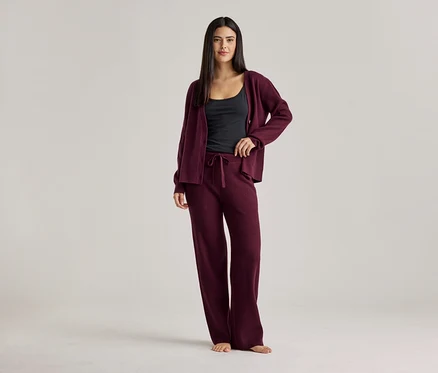 Une femme portant un débardeur noir, un cardigan bordeaux et un pantalon bordeaux. Elle se tient pieds nus sur un fond clair.