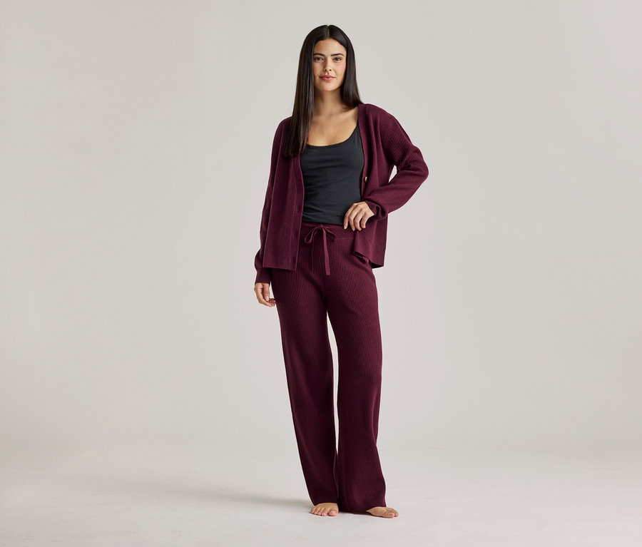 Une femme portant un débardeur noir, un cardigan bordeaux et un pantalon bordeaux. Elle se tient pieds nus sur un fond clair.