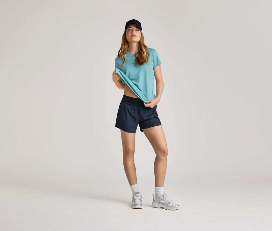 Une femme portant une casquette bleu marine, un t-shirt turquoise et un short bleu marine. Elle porte des chaussettes blanches et des baskets argentées.