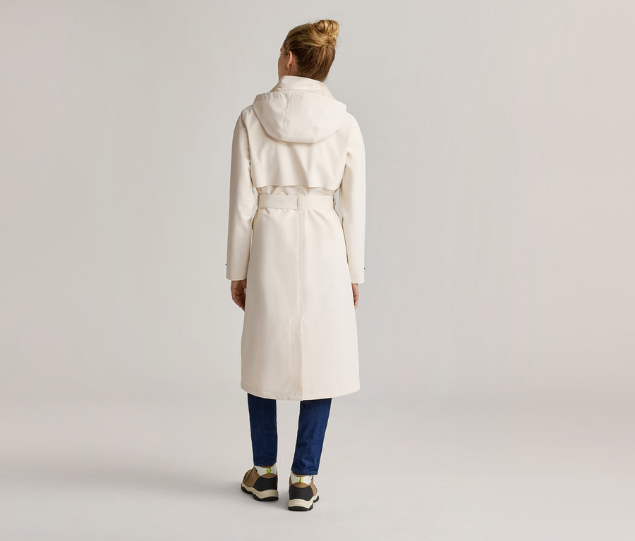 Vue arrière d'une femme portant un chignon, un long trench-coat clair à capuche et ceinture, un jean bleu et des baskets.