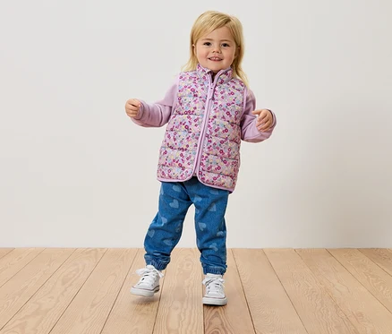 Une petite fille blonde se tient sur un plancher en bois, portant un gilet à fleurs, un haut violet et un jean.