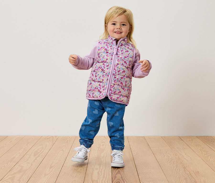 Une petite fille blonde se tient sur un plancher en bois, portant un gilet à fleurs, un haut violet et un jean.