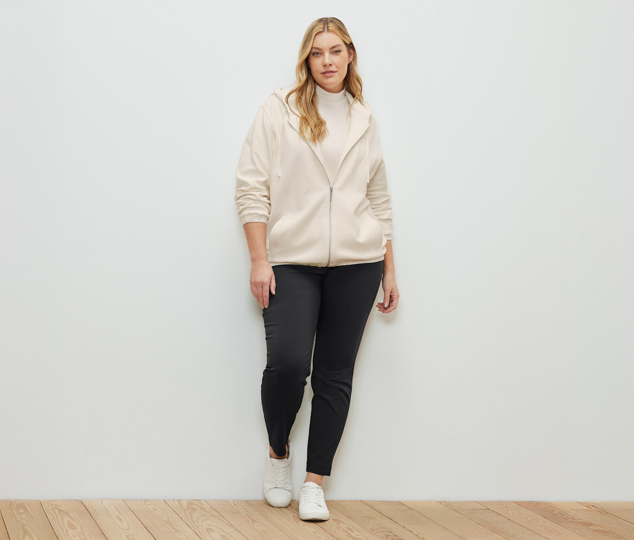 Une femme debout portant un sweat zippé couleur crème, un chemisier à col montant couleur crème et un pantalon stretch.