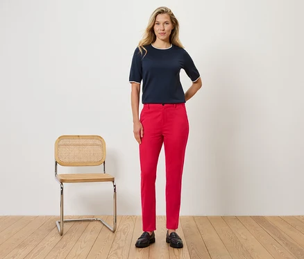 Une femme se tient devant un mur à côté d'une chaise. Elle porte un chemisier bleu marine avec des détails blancs et un pantalon rouge.
