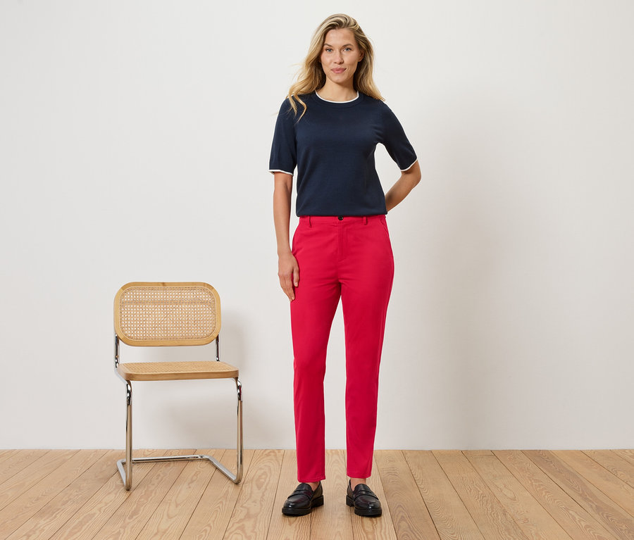 Une femme se tient devant un mur à côté d'une chaise. Elle porte un chemisier bleu marine avec des détails blancs et un pantalon rouge.