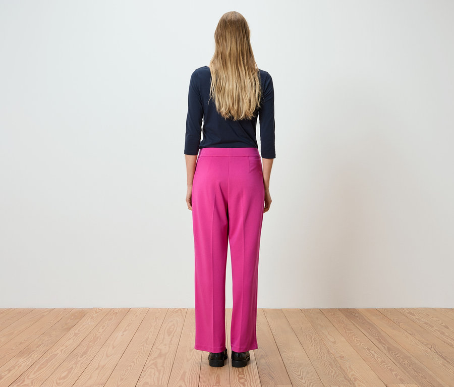 Vue de dos d'une femme aux longs cheveux blonds portant un t-shirt marine à manches longues et un pantalon rose.