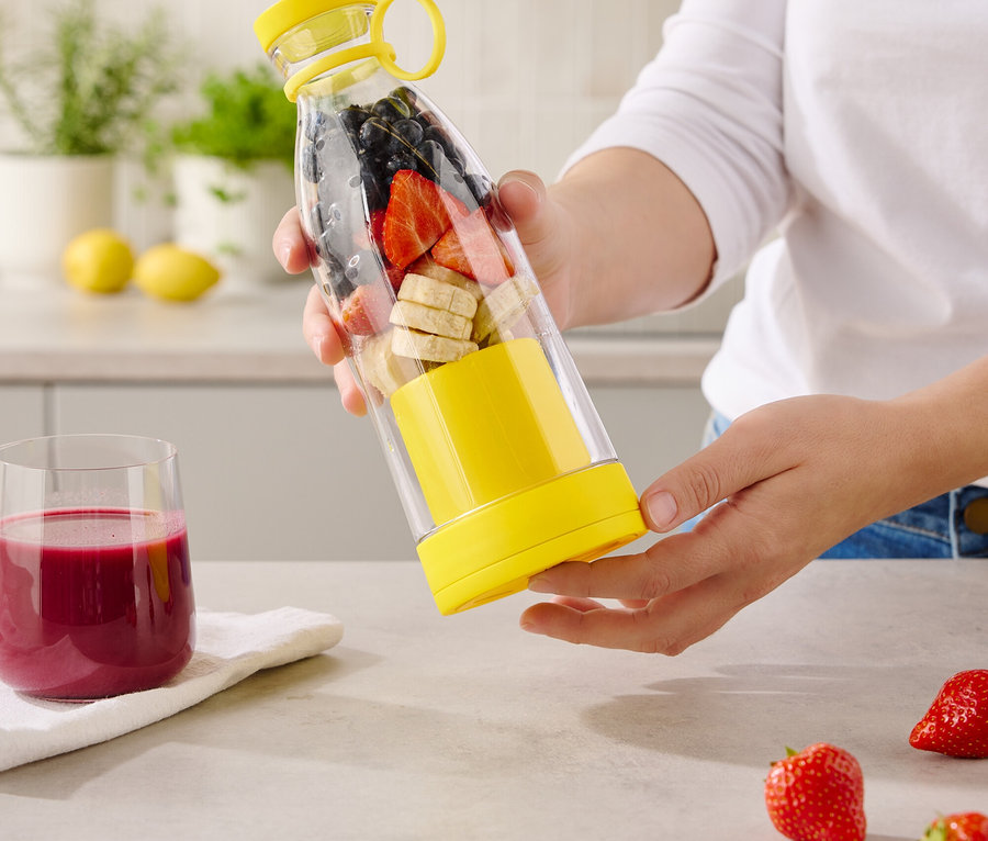 Une femme tient une bouteille transparente avec des accents jaunes, remplie de bananes, de fraises et de myrtilles.