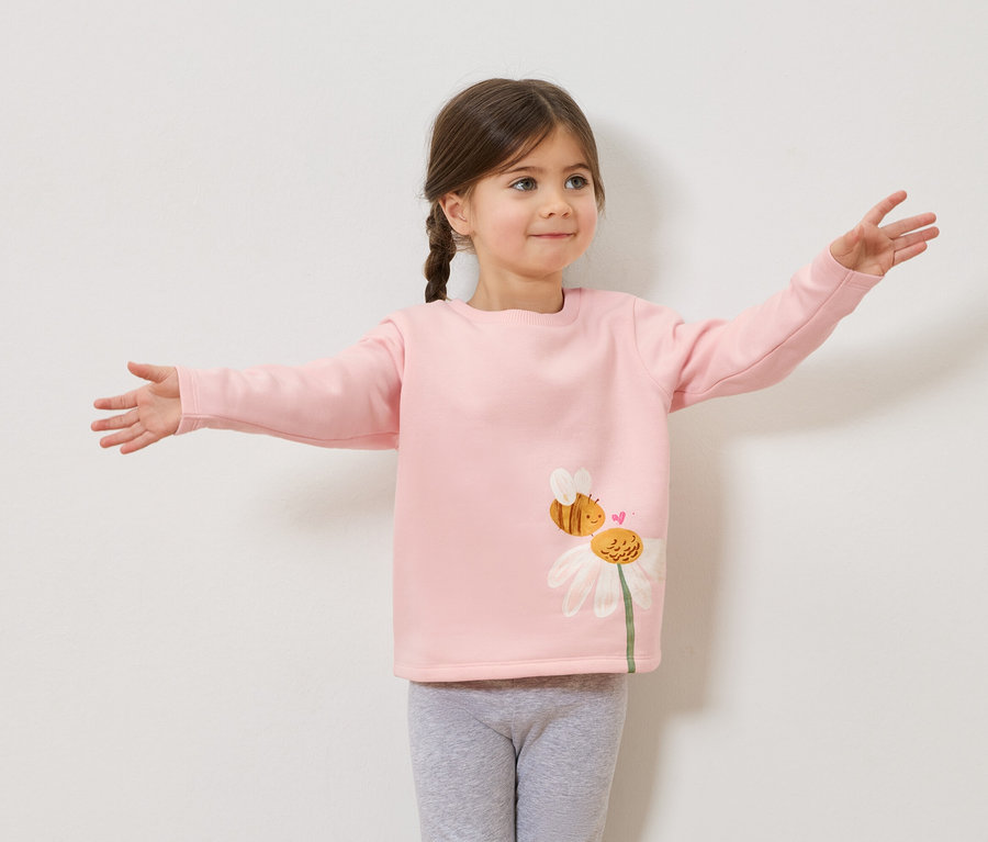 Fillette avec des tresses portant un pull rose avec un imprimé d'abeille et de marguerite, leggings gris, les bras écartés.