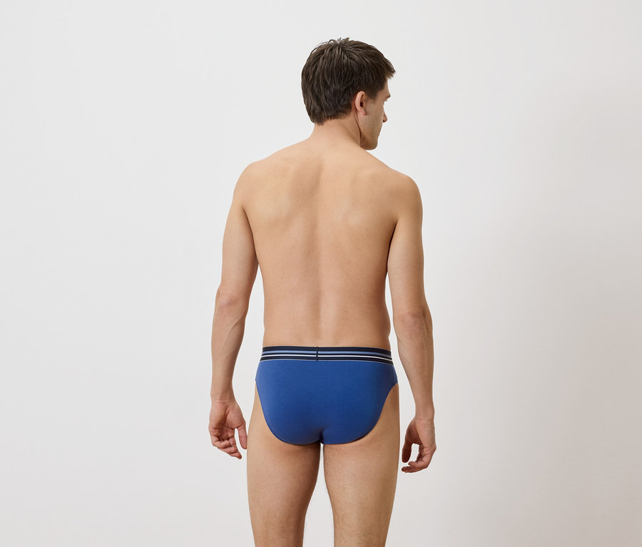 Vue de dos d'un homme portant un slip bleu avec une ceinture à rayures bleues et blanches sur un fond uni.
