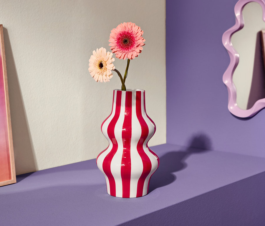 Deux fleurs de gerbera roses dans un vase rayé rouge et blanc sur une étagère violette, à côté d'une image rose et d'un miroir incurvé.