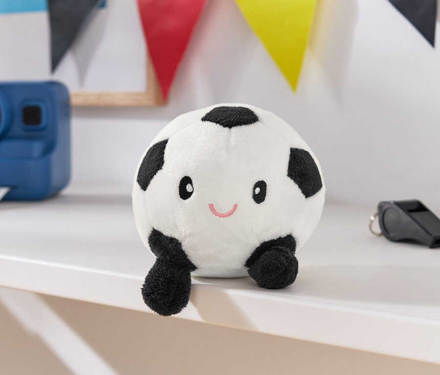 Un ballon de foot en peluche blanc avec des taches noires, un visage et de petites jambes est posé sur une surface blanche, à côté d'un appareil photo bleu et d'un sifflet noir.