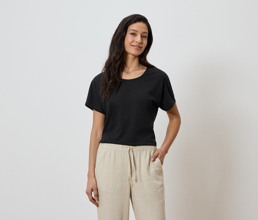 Femme portant un t-shirt noir et un pantalon beige, la main dans la poche.