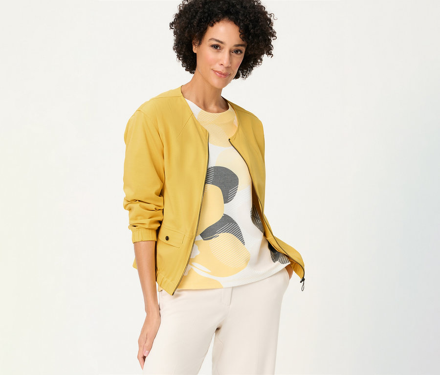 Une femme aux cheveux bouclés porte une veste en sweat Olsen jaune sur un haut à motifs et un pantalon clair.