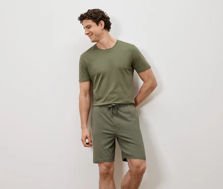 Un homme est appuyé contre un mur blanc, portant un t-shirt vert et un short.