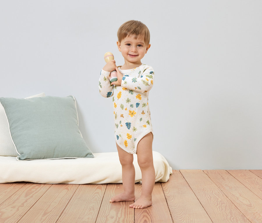 Un garçon souriant en body à motifs de feuilles tient un hochet. Un coussin et un futon sont visibles en arrière-plan.