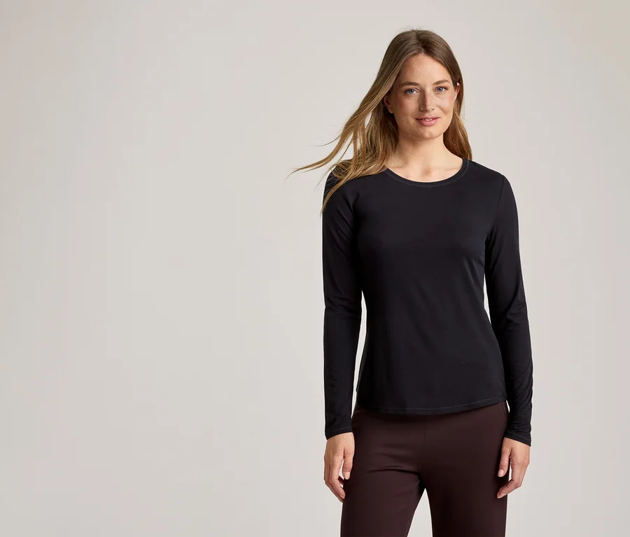 Une femme aux longs cheveux blonds ondulés porte un haut noir à manches longues et un pantalon marron châtaigne sur un fond gris clair.