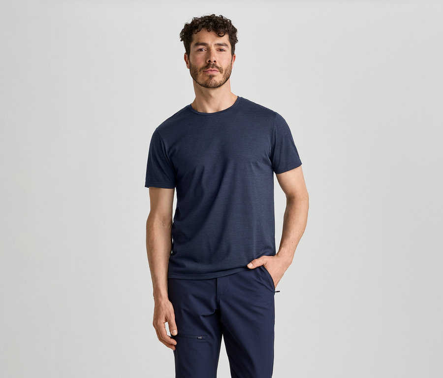 Un homme aux cheveux bouclés et barbu porte un t-shirt bleu foncé et un pantalon bleu foncé. Il se tient la main droite dans la poche.