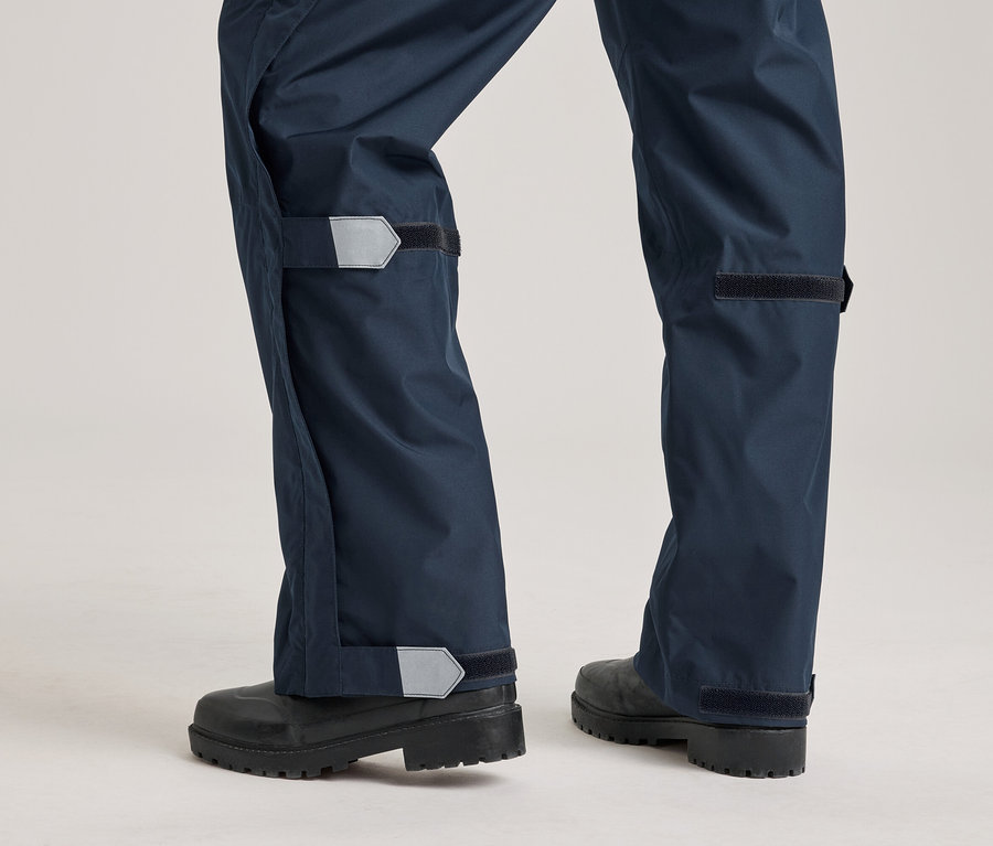 Gros plan d'un pantalon de pluie bleu marine avec fermeture velcro et détails réfléchissants, porté sur des bottes noires.