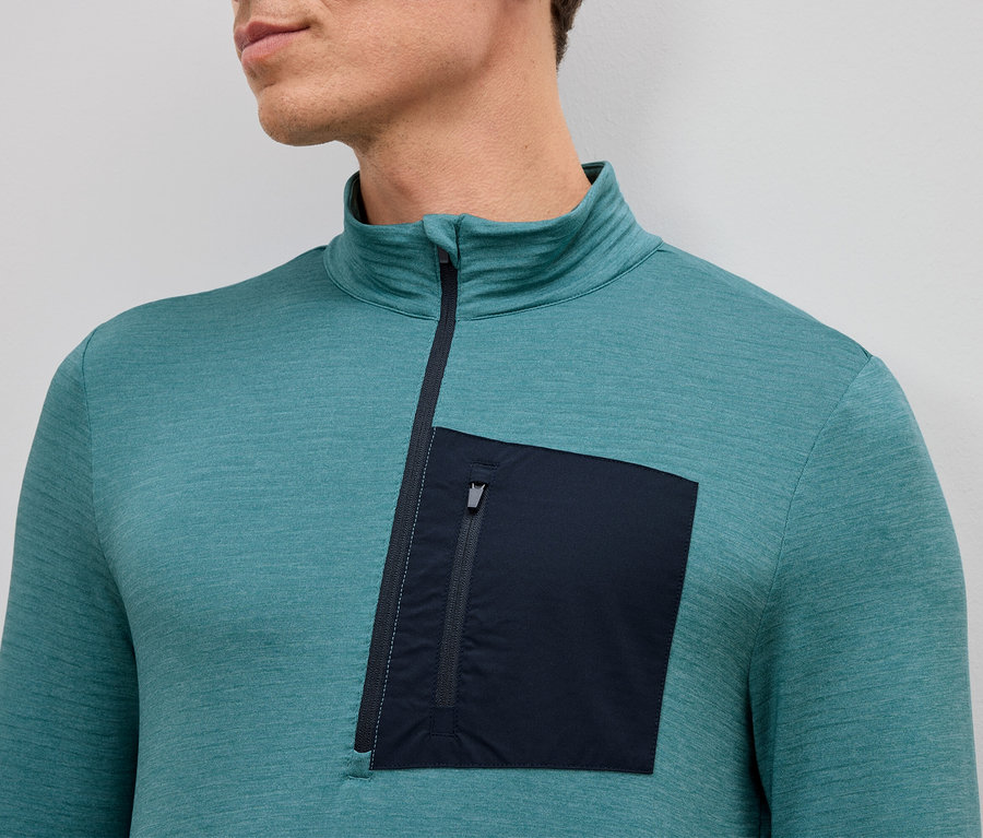 Gros plan d'un homme portant un T-shirt turquoise à manches longues avec une courte fermeture éclair et une seule poche plaquée zippée sur le côté gauche.