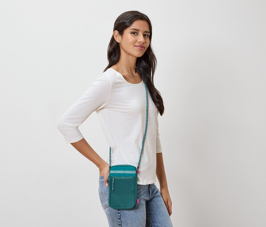 Une femme aux cheveux bruns foncés porte un sac à bandoulière vert avec une bandoulière turquoise, un t-shirt blanc à manches longues et un jean.