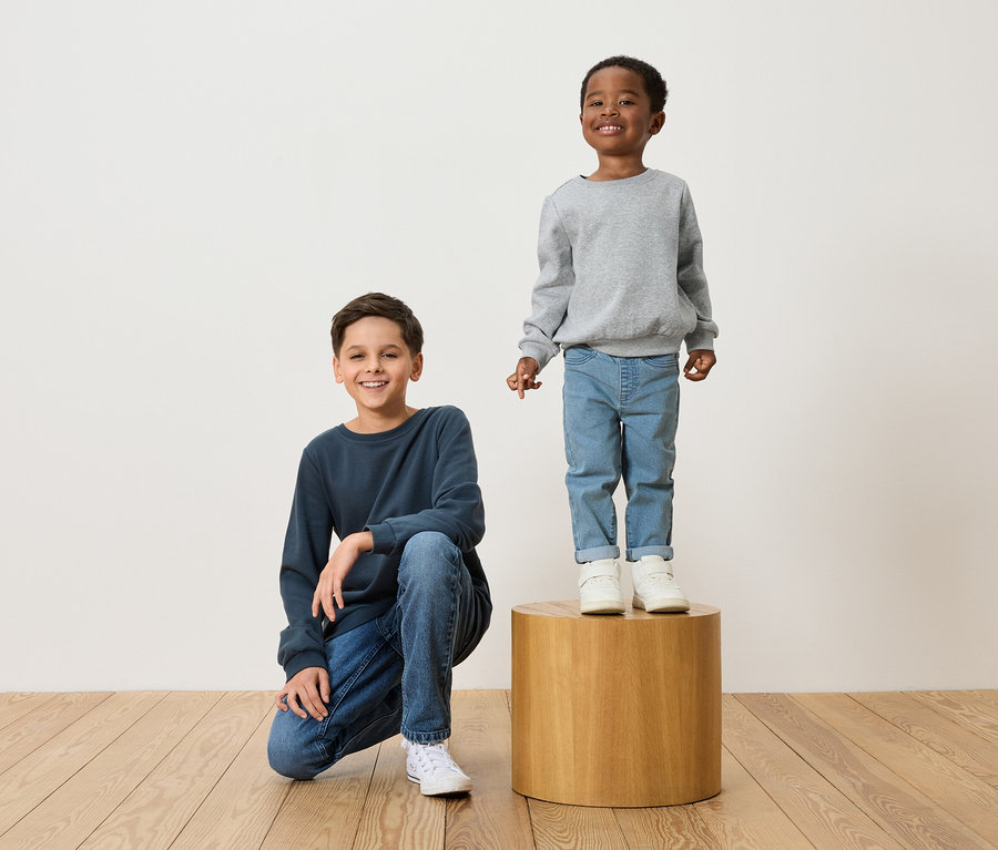 Deux garçons posent : l’un est accroupi en pull bleu marine et jean, l’autre se tient debout sur un tabouret en bois en pull gris et jean.