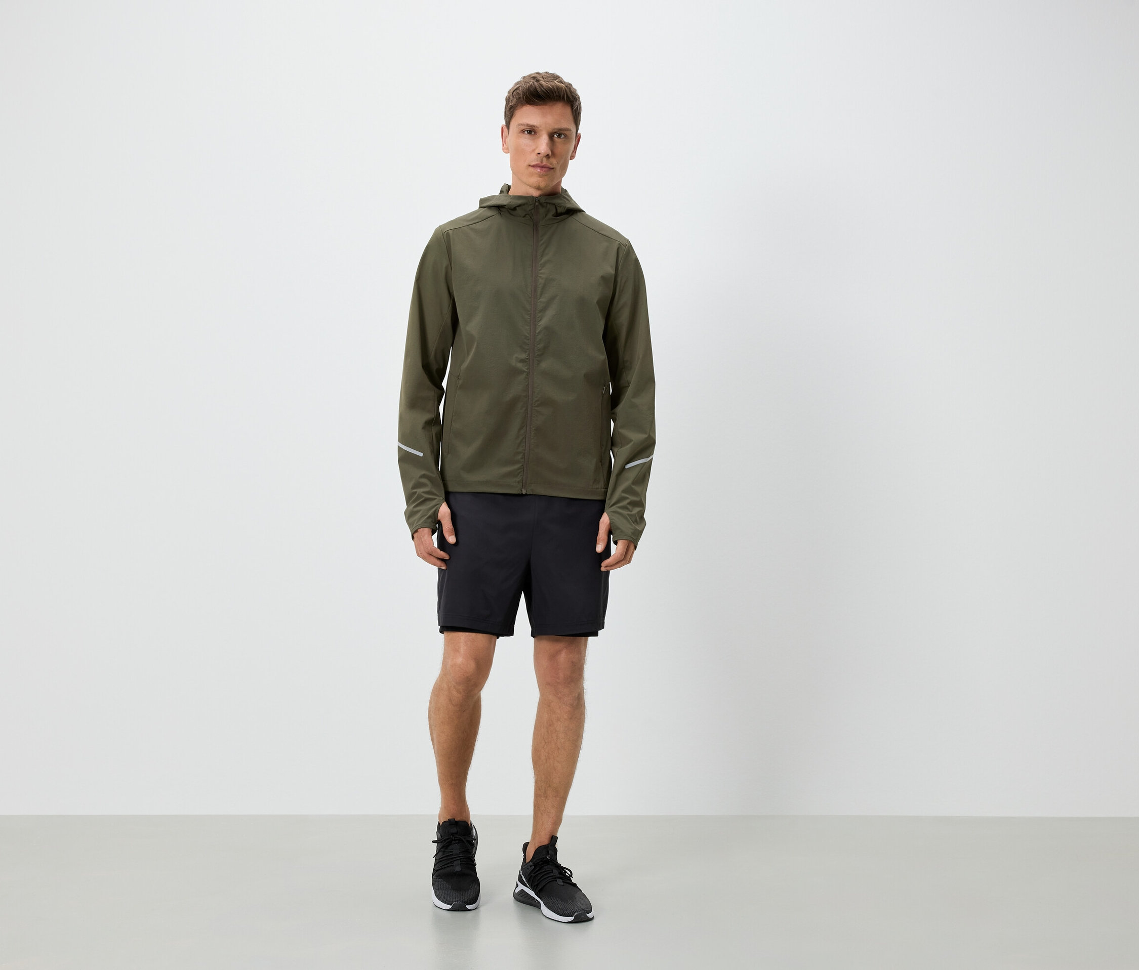 Homme portant une veste à capuche vert olive, un short noir et des baskets.